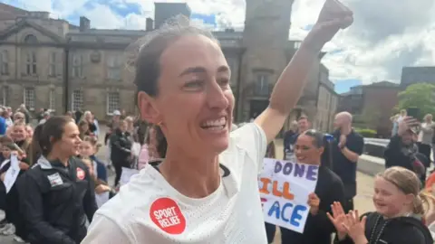 Jill Scott fist pumps the air as she makes her way through Keel Square in Sunderland. She is wearing a white t-shirt with a red sticker on it. There are crowds of people behind her cheering her on with signs. 