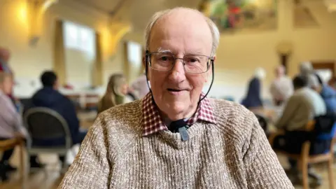 A older man in a brown jumper and brown shirt with spectacles smiling at the camera