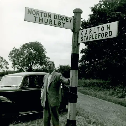 The Walt Disney Family Museum, San Francisco Black and white photo of Walt Disney in a loose suit touching the pole and looking up at the sign for Norton Disney with a black car parked behind him