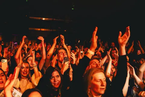 Getty Images A view of the crowd during a Myles Smith show - the crowd's hands are in the air and people are smiling, while illuminated dark orange from the stage lights.