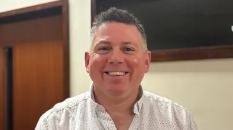 Danny O'hEarchai is a man with short grey hair. He is wearing a white shirt and smiling at the camera. A church hall is out-of-focus behind him.