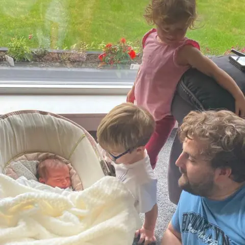 Lucy Lintott Smith Baby SS lies in a moses basket next to a window with a view to a garden. Her dad Tommy, wearing a blue t-shirt, and brother and sister are next to her. 