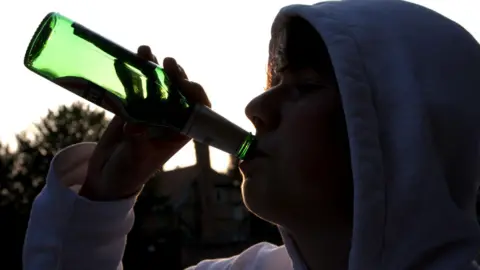 A boy wearing a blue hoodie swigs from a green beer bottle. He is outdoors and it is dusk.
