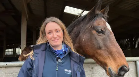 Emma Carter is standing next to a brown horse. Emma is wearing a blue navy jacket and has long brown hair. To the right is a brown horse with a white spot on its head. Its ears are pointing forwards.