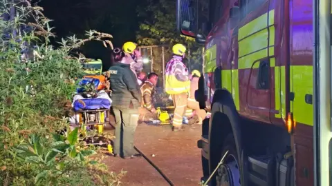 Harpenden Fire Station A paramedic with an ambulance bed stood alongside five firefighters by the front of a fire engine. Three of the firefighters are knelt on the ground. The paramedic looks on.
