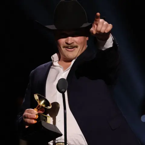Reuters Zach Top accepting his Grammy, holding the trophy in one hand, with his black cowboy hat obscuring his eyes, and pointing in the air with the other hand