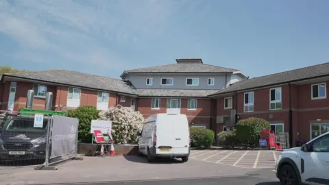 Picture of a brick built hospital in daylight with three sections connected together. There are a number of vehicles parked in front. 