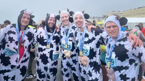 Josh McLaughlin/BBC Five men dressed in cow costumes during the Brighton Half Marathon 2026. They were medals around their necks which rest above the udders on their stomachs.