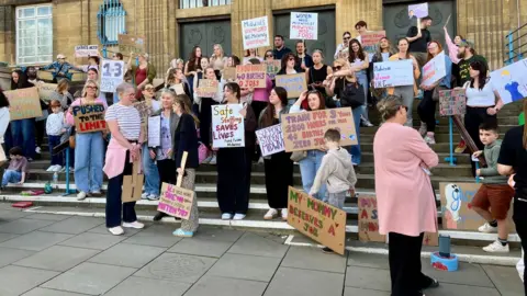 Nikki Fox/BBC Protesters stand in front of city hall in Norwich with posters and placards. One is held by a child and reads "my mummy deserves a job". Another says "pushed to the limit". Another reads "women need midwives, midwives need jobs." There are around 50 people in the photo and it shows around half of the protesters in total. They are standing on the concrete steps of city hall with the old building in the background.