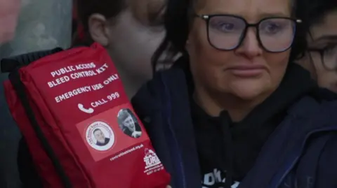 BBC A woman wearing black-rimmed glasses holds a red package marked "bleed control" kit and which also bears a picture of Harvey Willgoose