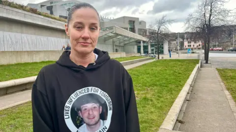 Lisa Petrie standing outdoors near a modern concrete building and grassy area, wearing a black hoodie with a circular graphic and text on the front. Pathways, trees without leaves, and cloudy skies are visible in the background