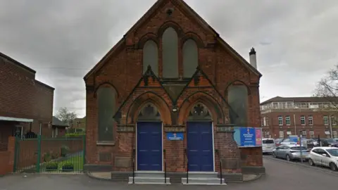 Google A small church with symmetrical blue doors and pointed arches at the front. There are several cars parked alongside the church on the road. 
