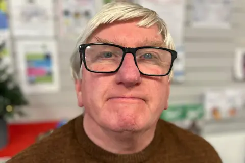 Gordon has white hair and is wearing thick-rimmed black glasses. He is wearing a brown jumper. 