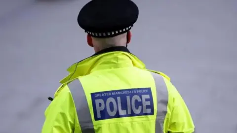 A rear view of a Greater Manchester Police officer wearing his uniform including a police hat and hi-vis jacket with the words 'Greater Manchester Police' on it.