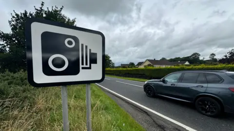 A speed camera sign on the side of a road with a car passing by