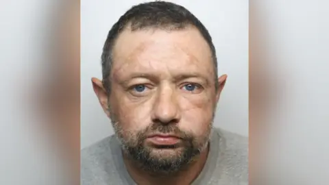 Stanley Saxon mug shot. He has short dark hair, blue eyes and a brown beard.