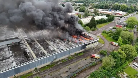 Aerial view of the fire at the former Delphi Diesel Systems site, showing thick black smoke pouring into the air while firefighters try to hose the site down.