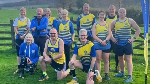 12 runners in yellow and blue striped running kits. They are a range of ages seemingly between about 40 and 70. They are all smiling at the camera and in a rural location, with a field behind them.