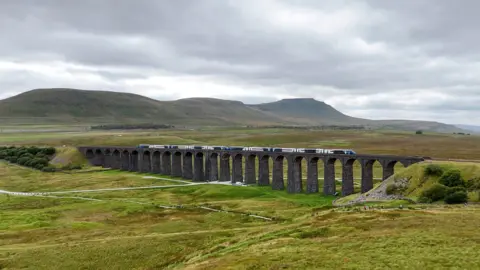 A train running along the Settle to Carlisle railway line over the Ribblehead Viaduct. The stone viaduct with its many arches is surrounded by rolling green land with fells in the background.