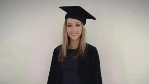 Family photo Jessica Gallagher smiling at the camera on her graduation day. She is wearing a black mortarboard and a black robe over a black or dark navy dress. She has long, straight blonde hair and is standing in front of a plain, pale background.