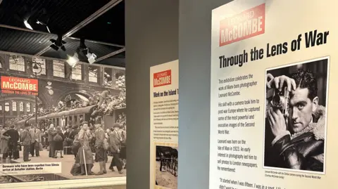LEONARD MCCOMBE A board with a picture of McCombe holding up his camera with the words Through the Lens of War above it, with a view through to the exhibition on the right.