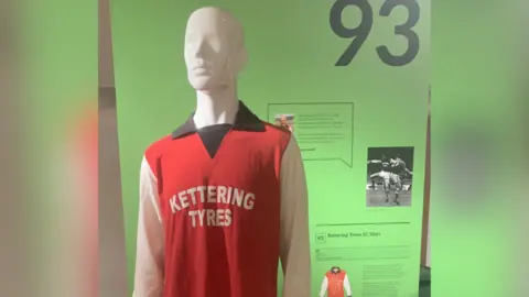 Bryan Lewin A mannequin on display is wearing the 1976 Kettering Town football kit. It is red with a black collar, white sleeves and "Kettering Tyres" printed in white font in the centre. Behind the mannequin is a green display outlining the history of the shirt.