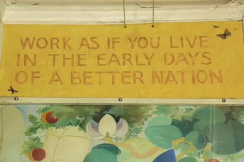 Alasdair Gray Painted mural detail from the Ubiquitous Chip’s Arcadia, featuring a yellow banner with red lettering that reads WORK AS IF YOU LIVE IN THE EARLY DAYS OF A BETTER NATION, accompanied by two small painted butterflies and a garden‑themed scene beneath it.