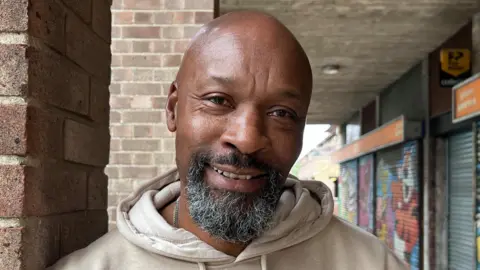Lily-May Symonds/BBC Albert Bernard is in the middle of the frame. He is looking at the camera smiling and is wearing a beige jumper. He is stood next to a brick pillar, and behind him, is a row of shop units which have their metal shutters down.