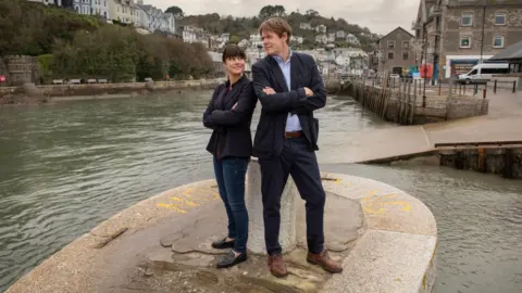 Red Planet Pictures Zahra Ahmadi and Kris Marshall are standing on the end of a pier surrounded with water in Looe. They have their backs turned against each other and their arms are folded as they turn and smile at each other. Behind them are granite shorefront houses and a harbour wall. 