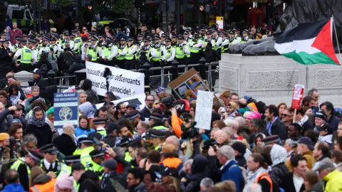 Reuters A crowd of people in Trafalgar Square protesting. A number of police in fluorescent jackets are among them and a Palestine flag is flying near one of the lion statues. In the background are a large number of police behind a fence