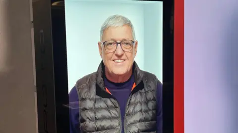 Hologram version of a man called Bill on a large screen, wearing a purple jumper and black gilet and smiling