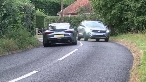 Two grey cars meeting at a sharp bend in a country road. They're each on their own side of the road but there is not much space between them. There's shrubbery on both sides of the road and a brick building in the background.