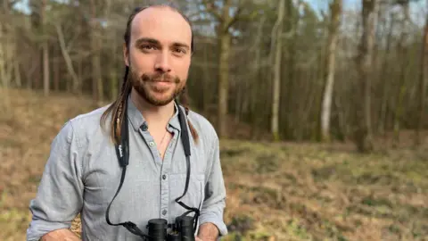 Fiona Irving / BBC A man with long tied-back hair and a beard looks at the camera. He is wearing a grey shirt and has a pair of binoculars around his neck. In the background is a woodland.