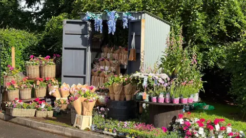 Jamie Kelly Flowers hang from, fill and surround a corrugated metal shed. Behind it there are green trees and shrubbery. 