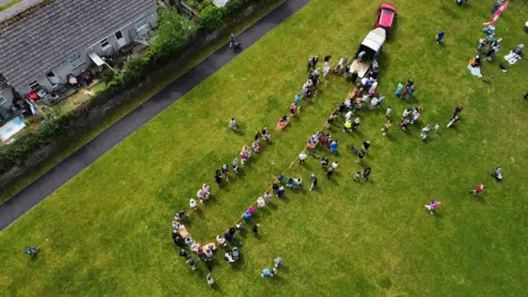 Mark Grice Aerial view of a large group of people standing in a winding line on a grassy field. A red vehicle with an open trailer is parked in the top right corner, with several people nearby. In the background, a row of houses with backyards lines the edge of the field, and a paved path runs parallel to the houses with a few individuals walking along it.