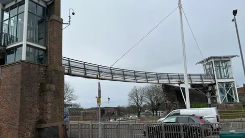 Irish Gate Bridge, also known as Millennium Bridge, in Carlisle. The large steel structure crosses a busy dual carriageway with traffic moving along the road beneath it. There are towers on either side containing lifts which have been permanently closed due to vandalism. The bridge has a slight curve to it and a tall white steel pole with cable attachments towards the far side.