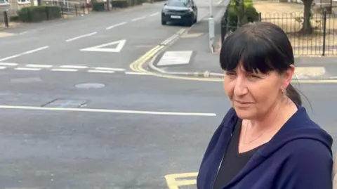 A woman in a navy hoodie with black hair is stood by a crossroads. In the middle of the crossroads is a manhole with a blue cross spray painted on it.
