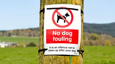 A sign saying No Dog Fouling is tied to a wooden post, underneath it reminds people it is an offence not to clean up after your dog