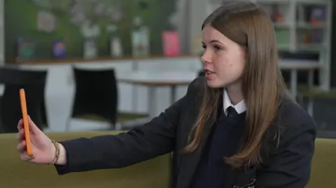 Imogen pictured sitting at her school, Appleton Academy in Bradford. She has long brown hair and is wearing a school blazer, jumper and white shirt. She is holding a green mobile phone out in front of her.