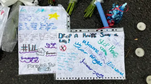 PA Media Pieces of white paper, inside plastic ring binder sheets, carry messages such as 'drop a knife, save a life' and are placed alongside flowers and candles.