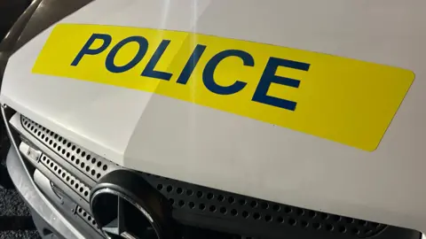The front of a police vehicle. The vehicle is white and it has the word 'Police' in a yellow box.