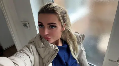A young blonde woman in a selfie. She wears a blue top and beige coat and has thick dark eyelashes.