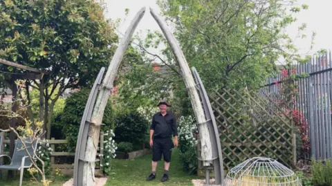 Lara King/BBC A man wearing a black shirt and shorts and a brown hat is standing underneath an archway which is made of the 134 year old jawbones of a whale. There are trees and plants surrounding the arch which is held up with steel supports. 