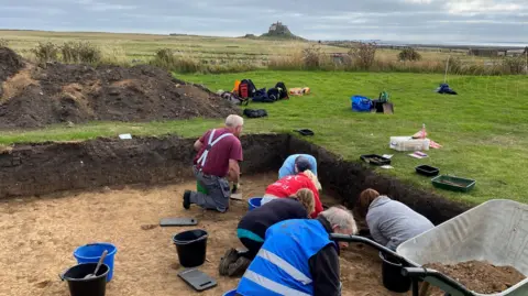 An archaeological excavation with a number of people kneeling down in a square pit. There is a wheelbarrow in the foreground and Lindisfarne can be seen in the distance.