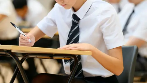 Student in school uniform writing on paper at a desk in a classroom, with other students blurred in the background.