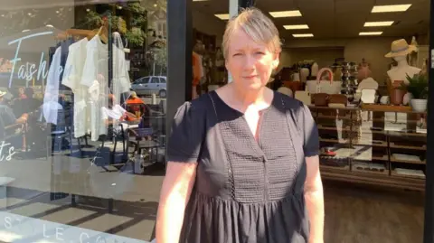 A woman with short blonde hair and wearing a brown top is smiling while standing outside the entrance to her shop. Items including clothing and hats can be seen on display inside.