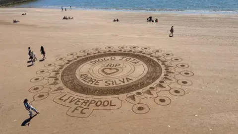 A large and intricate sand art design on a sandy beach. The artwork has multiple concentric circles and detailed patterns with the writing RIP Diogo Jota and Andre Silva at the centre of the artwork. There are some people along with the artist admiring it. 