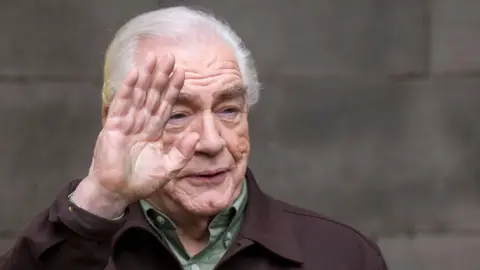 PA Media Brian Cox with white hair raising his right palm in a wave. He is only visible from the shoulders up and is wearing a light green shirt under a dark brown jacket.