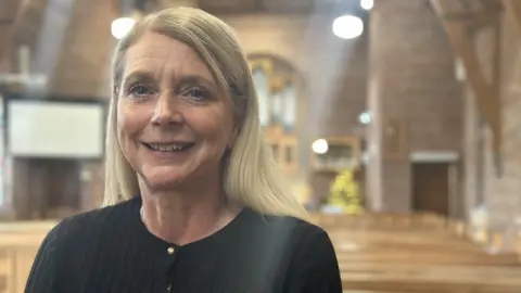 BBC A woman with blonde hair, smiles at the camera. She wears a black cardigan. Behind her the blurry outline of the inside of a church can be seen 