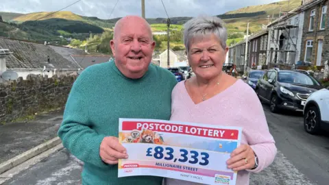 Alan and Muriel Owen smiling at the camera, they are holding their winning Postcode Lottery cheque. Alan is wearing a green jumper and is bald. Muriel is wearing a pink jumper and has short grey hair.